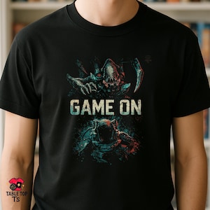 May include: Black t-shirt with the text "GAME ON" and a graphic design. The design features a detailed illustration of a creature and a futuristic figure in blue, red, and green. The shirt is from Table Top TS.