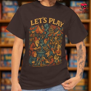以下が含まれることがあります： 「LET'S PLAY」の黄色い文字がプリントされた茶色のTシャツ。中世の村のカラフルなイラストが描かれており、人々が鍋や市場の屋台の周りに集まっている様子が描かれています。ヴィンテージでファンタジーなテーマです。