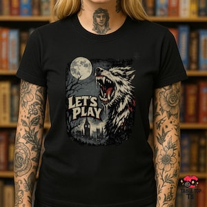 Puede incluir: Camiseta negra con un diseño de un lobo aullando bajo la luna llena. El diseño incluye el texto "LET'S PLAY" y una silueta de castillo espeluznante. El lobo tiene dientes afilados y detalles rojos.