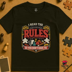 Peut inclure: T-shirt noir avec l'inscription "I READ THE RULES SO YOU DON'T HAVE TO". Le graphisme comprend des dés, des pions et le mot "RULES" en rouge. Le t-shirt est de Table Top TS.