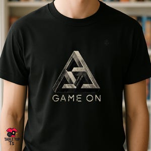 Könnte beinhalten: Schwarzes T-Shirt mit einem geometrischen Dreieck-Design und dem Text "GAME ON" in Weiß. Die Grafik hat einen 3D-Effekt, der eine optische Täuschung erzeugt. Das Shirt hat einen Rundhalsausschnitt.