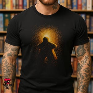 May include: A black short-sleeved t-shirt featuring a central graphic of a board game meeple silhouette. The meeple is rendered in a vibrant, textured orange and yellow light effect, appearing as if it is glowing against the dark fabric background.