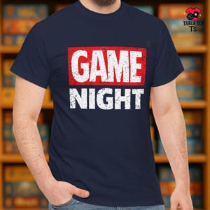 May include: A navy blue t-shirt featuring the text "GAME NIGHT" in a distressed white font, within a red rectangular frame. The shirt has a crew neck and short sleeves. The design is centrally placed on the chest.