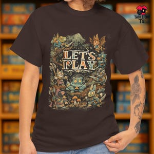May include: A brown t-shirt with a fantasy illustration and the words "LET'S PLAY". The design features cartoon characters, mushrooms, and a mountain scene. The shirt has a crew neck and short sleeves.