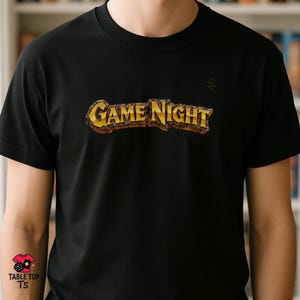 May include: Black t-shirt featuring the text "GAME NIGHT" in a gold and brown textured font. The shirt has a small logo on the upper right chest and a logo on the lower left. A casual wear item.