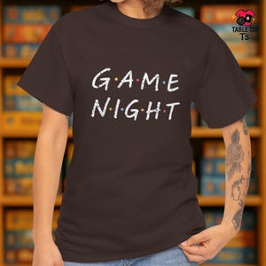 May include: A dark brown t-shirt featuring the text "GAME NIGHT" in white, with coloured dots above the letters. This casual top is suitable for game nights or relaxed social events.