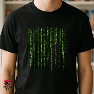 May include: A black short-sleeved t-shirt featuring a central graphic design composed of vertical columns of small green symbols that resemble board game meeples, arranged in a pattern reminiscent of falling digital code on a dark background.