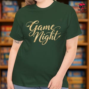 May include: A dark green t-shirt featuring the words "Game Night" in gold glitter script. The shirt has a crew neck and short sleeves. The brand logo "Table Top Ts" is in the upper right corner.