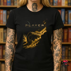 Pode incluir: Uma t-shirt preta de manga curta com a palavra PLAYER em texto dourado sobre uma ilustração gráfica de uma criatura circular grande e escura que emerge de uma paisagem dourada e rodopiante com duas pequenas figuras em primeiro plano.