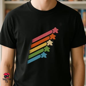Op de afbeelding: Zwart t-shirt met een kleurrijk grafisch ontwerp. Het ontwerp toont zes mensvormige spelstukken die op regenboogkleurige strepen omhoog gaan. De tekst "TABLE TOP TS" staat op een klein label linksonder.