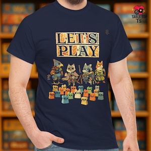 May include: Navy blue t-shirt with the text "LET'S PLAY" in a vintage style. The shirt features a graphic of anthropomorphic animal characters and game pieces, perfect for board game enthusiasts.