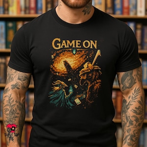 May include: Black t-shirt with a fantasy-themed graphic. The design features a wizard, a warrior, and the words "Game On" in gold. The artwork includes a cosmic background and card-like elements, suggesting a tabletop game theme.