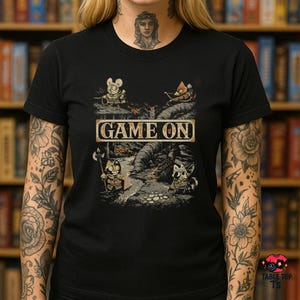 May include: Black t-shirt with the text "GAME ON" in a rectangular frame. The graphic shows cartoon characters in a fantasy setting, including a mouse, a fox, and two cat-like figures. The scene is set in a river.