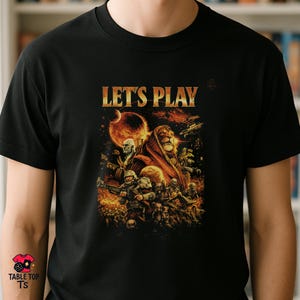 Puede incluir: Camiseta negra con un diseño gráfico dorado que dice "LET'S PLAY". El diseño incluye una figura parecida a un león, soldados y un planeta. La camiseta es de Table Top TS.