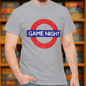 May include: A grey t-shirt with a red circle and a blue bar across it, displaying the words "GAME NIGHT" in white. The design resembles a London Underground sign. The shirt has a crew neck and short sleeves.