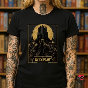 以下が含まれることがあります： 黒いTシャツ。金と黒のグラフィックで、城、人物、そして「LET'S PLAY」の文字が描かれています。デザインは、長方形の枠の中に、金色の月の背景に配置されています。