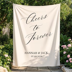 Op de afbeelding: Een crèmekleurige stoffen banner met de woorden "Cheers to Forever" in elegant zwart schrift. Daaronder staan de namen "Hannah & Jack" en de datum "07.05.2026" gedrukt. De banner wordt buiten gepresenteerd, omringd door groen.