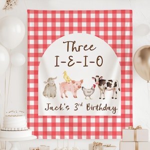 May include: A red and white checkered backdrop with the text "Three I-E-I-O" and "Jack's 3rd Birthday". The backdrop features illustrations of farm animals including a sheep, pig, duck, chicken, and cow. White and gold balloons are in the background.