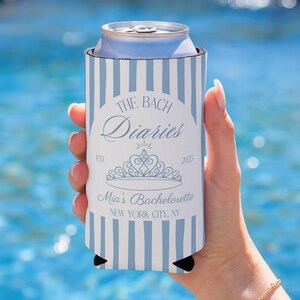 May include: A blue and white striped can cooler with the text "The Bach Diaries" and "Mia's Bachelorette" printed on it. The cooler has a tiara graphic and the year 2025. The words "New York City, NY" are also printed on the cooler.