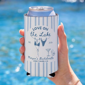 May include: A white and blue striped can cooler with the text "Love on the Lake" and "Morgan's Bachelorette Lake Tahoe, CA". The cooler features illustrations of swimwear, a cocktail, and a swan.