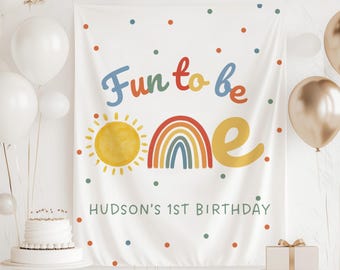 May include: White fabric banner with the text "Fun to be ONE" in colorful lettering, a yellow sun, and a rainbow. The banner also includes the text "HUDSON'S 1ST BIRTHDAY" in a lower font. The banner is decorated with colorful polka dots.