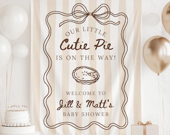 May include: A rectangular fabric banner featuring vertical beige and white stripes with a brown decorative border and bow. The text reads: OUR LITTLE Cutie Pie IS ON THE WAY! WELCOME TO Jill & Matt's BABY SHOWER, with a central pie illustration.