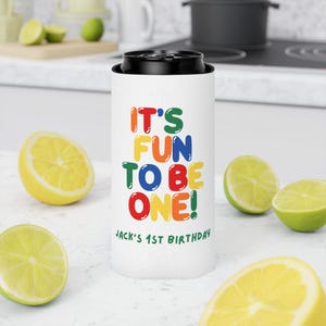 May include: A white can cooler with the text "IT'S FUN TO BE ONE!" in colorful letters. Below the text is "JACK'S 1ST BIRTHDAY" in black. The cooler is surrounded by cut lemons and limes.