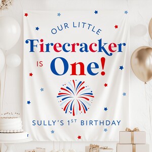 May include: A white rectangular fabric backdrop featuring the text "OUR LITTLE Firecracker IS One!" in blue and red lettering, with a central red, white, and blue firework graphic and the text "SULLY'S 1ST BIRTHDAY" at the bottom.