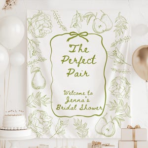 Peut inclure: Une bannière blanche avec des illustrations botaniques vertes et le texte "The Perfect Pair. Welcome to Jenna's Bridal Shower." La bannière est ornée de poires, de fleurs et de feuilles. Des ballons blancs et dorés et un gâteau sont également visibles.