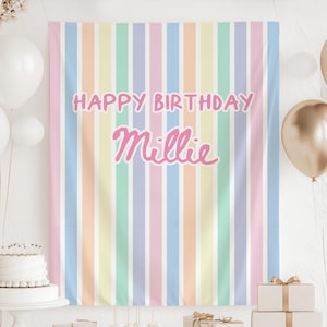 May include: A pastel-colored birthday backdrop with vertical stripes in shades of pink, blue, yellow, and green. The words "HAPPY BIRTHDAY Millie" are written in pink script across the center. White and gold balloons and a cake are in the background.