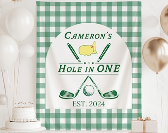 Op de afbeelding: Een witte banner met groene tekst "Cameron's Hole in One EST. 2024" met golfclub- en balafbeeldingen. De banner staat tegen een groen-witte geruite achtergrond, met ballonnen en een taart op de achtergrond.