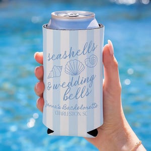 May include: A blue and white striped can cooler with the text "seashells & wedding bells" and seashell illustrations. The text "Jenna's Bachelorette, Charleston, SC" is also printed on the cooler. The cooler is holding a silver can.