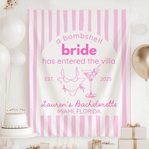 May include: A white banner with pink vertical stripes and text that reads "a bombshell bride has entered the villa." The banner includes a martini glass and bikini graphic, with "Lauren's Bachelorette, Miami, Florida" below.