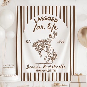 以下が含まれることがあります： 茶色の縦縞模様と「LASSOED for life」の文字が入った白いバナー。中央にはカウボーイのイラスト。バナーには「Jenna's Bachelorette, Nashville, TN」と「Est. 2026」の文字も入っています。