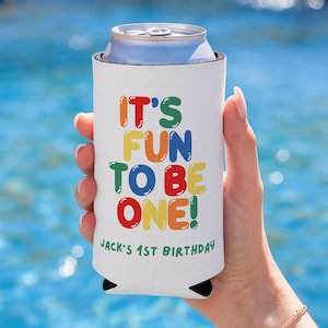 May include: White can cooler with the text "IT'S FUN TO BE ONE!" in colorful letters. The text "Jack's 1st Birthday" is printed below. The cooler is holding a silver can, against a blue water background.