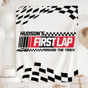 May include: White banner with black and red text that reads "HUDSON'S FIRST LAP AROUND THE TRACK." The banner has a race car graphic and a checkered flag design. Balloons and a cake are in the background.