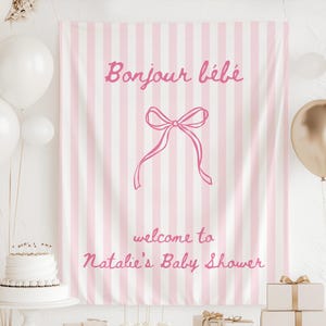 May include: A pink and white striped backdrop with the text "Bonjour bébé" and "welcome to Natalie's Baby Shower" in pink script. A pink bow graphic is centered on the backdrop. White and gold balloons and a white cake decorate the scene.