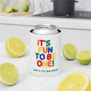 May include: White can cooler with the text "IT'S FUN TO BE ONE!" in colorful letters. The text "JACK'S 1ST BIRTHDAY" is printed below. The cooler is holding a silver can. Sliced lemons and limes are scattered around the cooler.