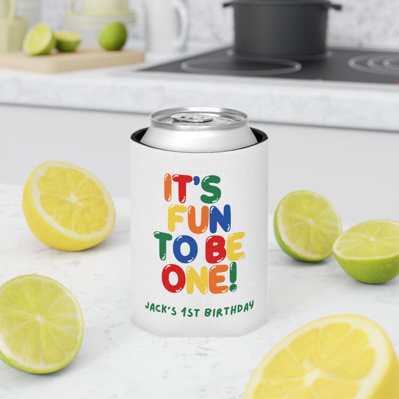 May include: White can cooler with the text "IT'S FUN TO BE ONE!" in colorful letters. The text "JACK'S 1ST BIRTHDAY" is printed below. The cooler is holding a silver can. Sliced lemons and limes are scattered around the cooler.