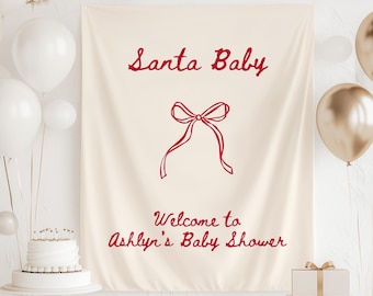 Peut inclure: Une bannière crème avec le texte rouge "Santa Baby" et "Welcome to Ashlyn's Baby Shower". Un nœud rouge est dessiné en dessous de la première ligne de texte. Des ballons blancs et dorés sont en arrière-plan.