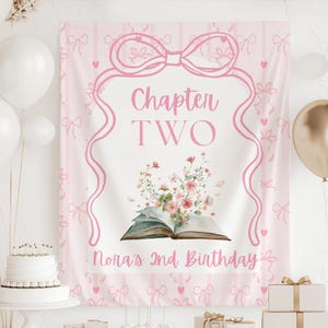 May include: A pink birthday banner with the text "Chapter TWO" and "Nora's 2nd Birthday". The banner features a floral illustration above an open book and is decorated with pink bows and ribbons. White and gold balloons are in the background.