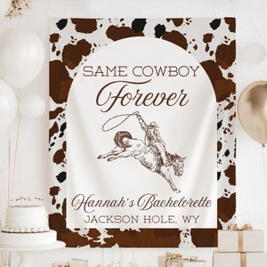 Può includere: Un banner bianco con bordo e sfondo con stampa maculata marrone. Lo striscione recita "Same Cowboy Forever" con un disegno di un cowboy su un cavallo selvaggio. È incluso anche il testo "Hannah's Bachelorette, Jackson Hole, WY".