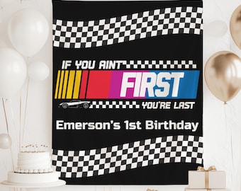May include: Black banner with the text "IF YOU AINT FIRST YOU'RE LAST" and "Emerson's 1st Birthday". The banner has a race car graphic and a checkered flag design. Balloons and a cake are in the background.