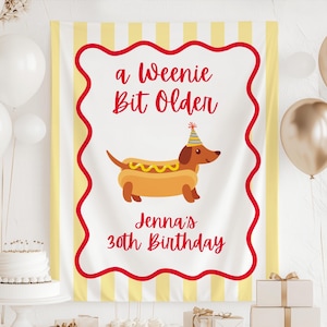 May include: A birthday banner featuring a cartoon dachshund in a hot dog bun wearing a party hat. The banner reads "a Weenie Bit Older" and "Jenna's 30th Birthday" in red script on a white background with a red border and yellow stripes.