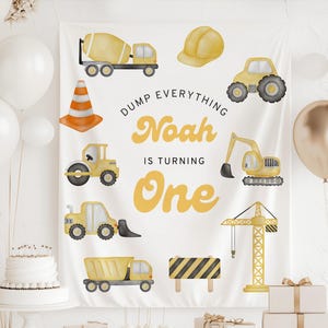 May include: A white banner with yellow construction vehicles and a hard hat. The banner reads "Dump Everything Noah is Turning One" in yellow lettering. Balloons and a cake are in the background.