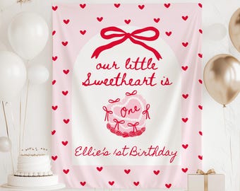 May include: A pink birthday banner with red hearts and text that reads "our little Sweetheart is One of Ellie's 1st Birthday." The banner has a large red bow and a heart-shaped cake design.