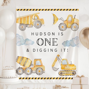 May include: A white banner with watercolor illustrations of construction vehicles, including a dump truck, excavator, and cement mixer. The text "HUDSON IS ONE & DIGGING IT!" is displayed in black. Yellow and black hazard stripes border the top and bottom.