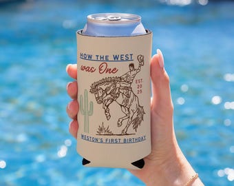 May include: A beige can cooler with a cowboy and bucking bronco illustration. Text reads "HOW THE WEST was One" and "WESTON'S FIRST BIRTHDAY". A cactus is also featured in the design. The cooler is held in front of a blue water background.