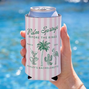 May include: A pink and white striped can cooler with the text "Palm Springs Before The Rings" and "Ashley's Bachelorette". The design includes illustrations of a cocktail, palm tree, cactus, and flamingo. The date "March 2026" is also printed on the cooler.