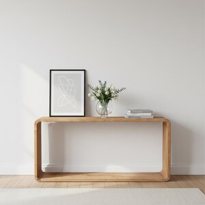 May include: A light oak console table with a curved design. It features a framed abstract print, a glass vase with white flowers, and two books. The background is a white wall and a light-coloured rug.